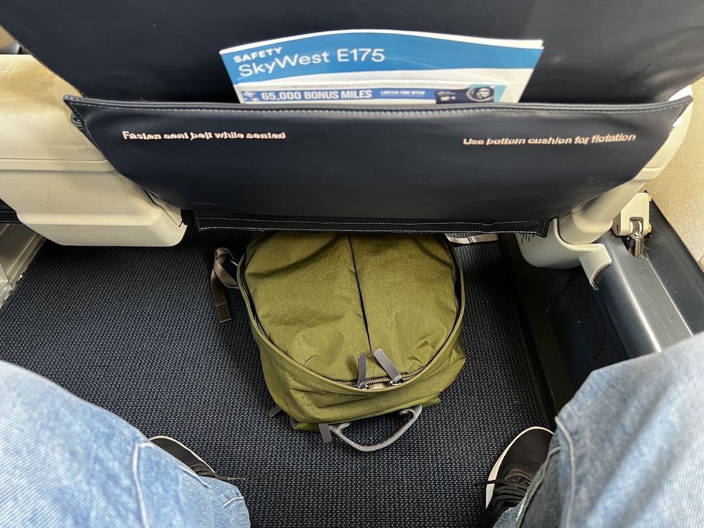 ERJ-175 일등석 좌석 밑 공간 — 올리브색 백팩이 빠듯하게 들어간 모습, 시트 포켓에 SkyWest E175 안전 카드